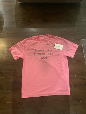 Rosa Acne Studios t-shirt Stockholm - Säljer en rosa t-shirt från Acne Studios med trycket 'Acne Studios STOCKHOLM 1996' på bröstet. Klassisk rund hals och korta ärmar. Perfekt för dig som gillar streetwear och vill ha något stilrent men ändå sticker ut.