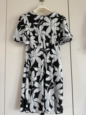 Svartvit blommig kortklänning - Kort klänning med svart botten och stora vita blommor. Puffiga korta ärmar och rund halsringning. Perfekt för dig som gillar ett statement-mönster. Materialet känns lätt och luftigt. Upplevs liten i storleken. 
