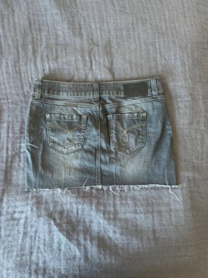 Grå jeanskjol  - Säljer en grå jeanskjol i kort modell med rå, fransig kant nedtill. Kjolen har klassiska fem fickor, bälteshällor och knappstängning framtill. Perfekt för dig som gillar en avslappnad och trendig stil.