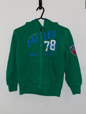 Grön hoodie med tryck och huva - Grön hoodie med dragkedja och huva. Tryck på bröstet med texten 'EAGLES 78' i blått och vitt samt ett märke på ena ärmen. Känguruficka framtill och ribbade muddar vid ärmslut och nederkant. Perfekt för en sportig look.