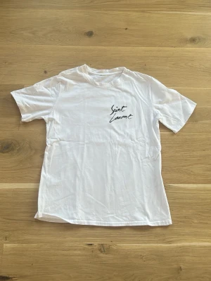 Vit Saint Laurent t-shirt - Vit t-shirt med Saint Laurent-tryck på bröstet. Klassisk rund hals och korta ärmar. Enkel och stilren design som passar till det mesta. Tillverkad i mjukt material för en bekväm känsla.