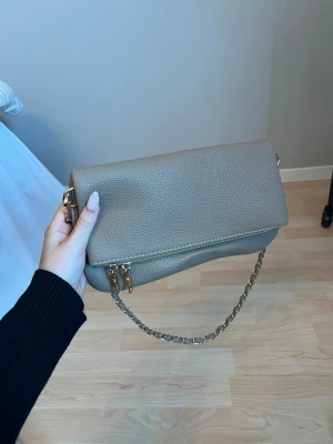 Väska - Stilren beige handväska i mjukt, strukturerat konstläder med viklock och guldfärgade detaljer. Längre band i väskan. Aldrig använd.