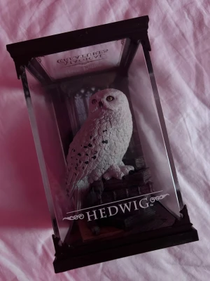 Harry Potter Hedwig prydnadsfigur - Säljer en detaljerad prydnadsfigur av ugglan Hedwig från Harry Potter, i vit och svart färg. Figuren står i en genomskinlig displaybox med svart ram och texten 'HEDWIG' på framsidan. Perfekt för samlare eller Harry Potter-fans.
