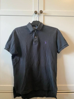 Svart pikétröja från Ralph Lauren - Klassisk svart pikétröja från Ralph Lauren med lila broderad logga på bröstet. Tröjan har korta ärmar, krage och knäppning med två knappar. Tillverkad i bomull för en bekväm och stilren look.