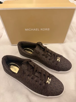 Bruna Michael Kors sneakers - Helt nya , dem är aldrig använda. Dem är köpta för 1700kr men jag säljer dem helt nya för 1250kr. Bruna sneakers från Michael Kors med klassiskt MK-mönster över hela skon. Storlek 40 men passar även mig som har strl 39. Tillverkade i ett tåligt material med lyxig känsla och stilren design. Har aldrig fått användning till dem, inga tecken på användning då dem är helt vita under skon och inga smutsiga fläckar eller något.