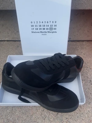 Maison Margiela svarta sneakers - Svarta sneakers från Maison Martin Margiela med en clean och stilren design. Skorna har en kombination av mocka och läderpaneler, svarta skosnören och en svart yttersula med logotyp under. Perfekta för dig som gillar minimalistisk och exklusiv stil.