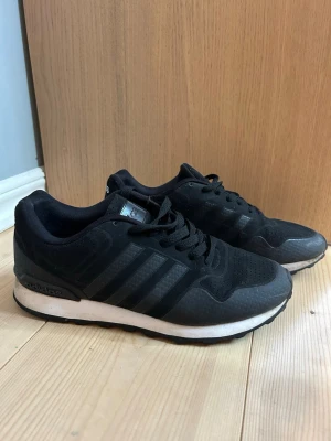 Svarta Adidas sneakers med vita sulor - Snygga svarta sneakers från Adidas med klassiska tre ränder på sidan och vit sula. Skorna har en låg profil och är tillverkade i en mix av textil och syntetmaterial. Perfekta för dig som gillar en clean och sportig look.