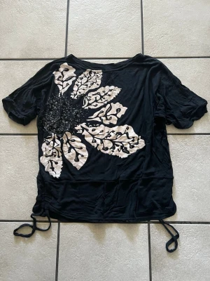 Svart t-shirt med stor blommig print storlek L/XL - Svart t-shirt med korta ärmar och stor vit blommig print framtill. T-shirten har snörning i sidorna för en cool detalj och avslappnad passform. Perfekt för dig som vill sticka ut lite extra med ett unikt tryck.