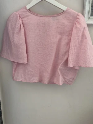 Ljusrosa blus med puffärmar - Supersöt ljusrosa blus med korta puffärmar och rund halsringning. Blusen har en liten öppning med knapp i nacken och är tillverkad i ett mjukt bomullstyg. Perfekt för dig som gillar en feminin och trendig stil.