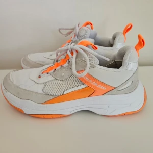 Sneakers från Calvin Klein Jeans - Chunky sneakers från Calvin Klein Jeans i vitt och ljusgrått med orange detaljer och meshpaneler. Lite smutsiga men fortfarande fint skick. Storlek 39 och jag tycker att dom är normala i storlek.