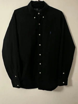 Ralph Lauren skjorta - Säljer en svart långärmad skjorta i manchester med klassisk krage och vita knappar framtill. Skjortan har en diskret broderad blå Ralph Lauren logga på bröstet och är perfekt för dig som gillar en stilren look. Haft på mig denna skjorta ett fåtal gånger under vissa speciella event, annars är den knappt använd och nästan i helt nytt skick. Hör av dig vid frågor eller funderingar.