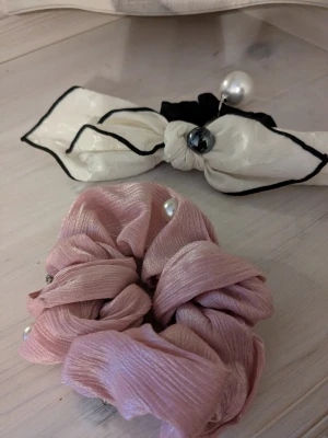Två håraccessoarer med pärldetaljer - Säljer två snygga håraccessoarer: en scrunchie i glansigt ljusrosa tyg med pärlor och en vit hårrosett med svarta kanter och pärldetaljer. Perfekta för att ge frisyren en extra touch och passar till många olika looks. Köpta i Kina 