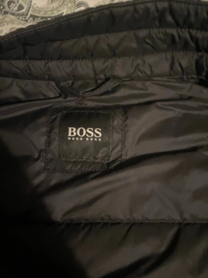 Svart pufferjacka från BOSS - Snygg svart pufferjacka från BOSS med klassisk quiltad design och diskret logotyp på bröstet. Jackan har en glansig yta och är tillverkad i ett slitstarkt syntetmaterial som håller värmen under kalla dagar. Perfekt för dig som vill ha en stilren vinterjacka.