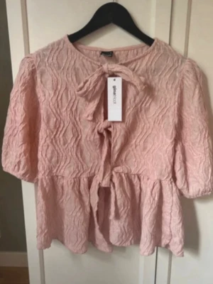 Rosa strukturerad blus med knyt - Supersöt ljusrosa blus från Gina Tricot med puffiga trekvartsärmar och trendig struktur i tyget. Blusen har knytband både vid halsen och midjan samt en lätt volang nedtill. Perfekt för en chic och avslappnad look.