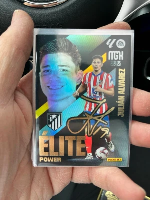 Panini fotbollskort Julián Alvarez - Samlarobjekt: Panini fotbollskort med Julián Alvarez i Atlético Madrids rödvitrandiga matchtröja. Kortet har texten 'Élite Power' och en tryckt autograf i guld. Baksidan har spelarinfo och statistik. Perfekt för fotbollsfans och samlare.