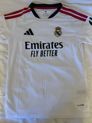 Real Madrid matchtröja Adidas vit - Snygg vit Real Madrid matchtröja från Adidas med svarta och röda detaljer. Tröjan har klubbmärke, Adidas-logga och Emirates Fly Better-tryck på bröstet. Tillverkad i lätt och ventilerande material med Climalite-teknologi. Perfekt för fotbollsträning eller match.