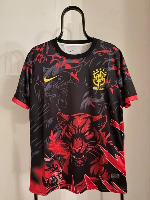 Nike Brasil svart/röd matchtröja - Snygg svart och röd matchtröja från Nike med Brasil-logga och gult swoosh på bröstet. Tröjan har ett stort tigertryck framtill, klösmärken bak och coola grafiska detaljer i rött och grått. Tillverkad i lätt, snabbtorkande material. Perfekt för fotboll eller streetwear.