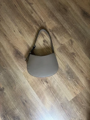 Beige axelremsväska i skinnimitation - Snygg beige handväska med axelrem och minimalistisk design. Väskan har en rundad form och justerbart spänne på remmen. Tillverkad i slät skinnimitation som ger en stilren look. Perfekt för dig som gillar enkel och trendig stil.