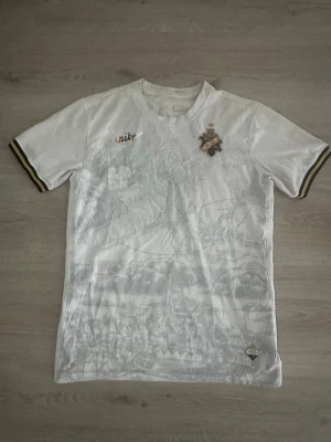 Vit AIK x Nike Stockholm Edition t-shirt - Exklusiv vit AIK x Nike Stockholm Edition t-shirt från 2023 med subtilt stadsmotiv i tyget. Kortärmad modell med gulddetaljer på ärmsluten och AIK-emblem på bröstet. Perfekt för dig som vill ha något unikt och sportigt från Nike.