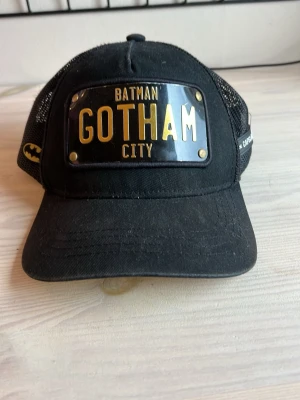 Svart Batman Gotham City keps - Snygg svart keps från Capslab med Batman-tema och texten 'Gotham City' i guld på en metallplatta framtill. Kepsen har mesh på sidorna och baksidan samt gul undersida på skärmen. Justerbar baktill för perfekt passform.