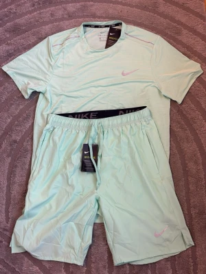 Nike mintgrönt träningsset - Snyggt träningsset från Nike i mintgrönt, består av en kortärmad t-shirt och matchande shorts. Båda plaggen har Nike-logga i ljusrosa och är tillverkade i ett lätt, andningsbart material som passar perfekt för träning och sportaktiviteter.