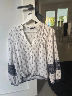 Vit blus med blått mönster Zara  - Luftig vit blus med blått bohemiskt mönster och dekorativa detaljer längs ärmslut och nederkant. Blusen har v-ringning, veckade axlar och långa ärmar med breda muddar. Perfekt till jeans eller kjol för en avslappnad men stilsäker look.
