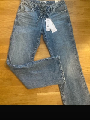 Blå lace-up jeans med raka ben - Säljer ett par blå jeans med snygg snörning framtill i midjan. Jeansen har raka ben och klassisk femficksdesign. Materialet är denim i en tvättad blå nyans som ger en avslappnad look. Perfekta för dig som vill ha något trendigt och bekvämt.