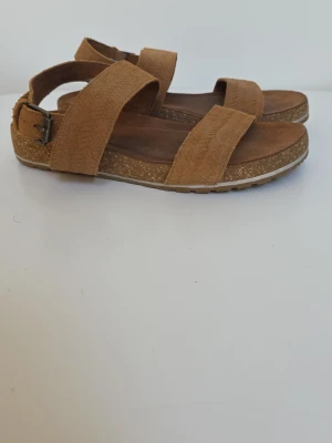 Bruna mockasandaler från Timberland - Snygga sandaler från Timberland i mjuk brun mocka med två breda remmar och justerbart spänne vid ankeln. Fotbädden är bekväm och sulan har en naturlig look med vita detaljer undertill. Perfekta för en avslappnad och stilren sommarstil. Storlek 39