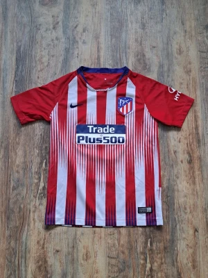 Atlético Madrid matchtröja Griezmann - Säljer en röd och vit randig Atlético Madrid matchtröja från Nike med Griezmann och nummer 7 på ryggen. Tröjan har klubbmärke, sponsortryck och La Liga-märke på ärmen. Tillverkad i lätt och ventilerande material, perfekt för fotbollsträning eller match.