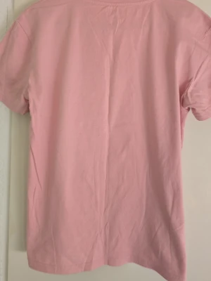 Rosa t-shirt med nalletryck - Söt rosa t-shirt med ett stort tryck av en nallebjörn och bokstäverna 'HI' på framsidan. T-shirten har klassisk passform och korta ärmar. Perfekt för dig som gillar gulliga motiv och vill ha en avslappnad stil.