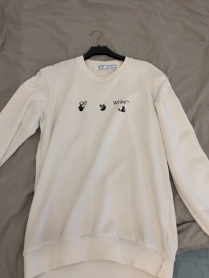Vit sweatshirt från Off-White - Vit sweatshirt från Off-White med svart tryck framtill och stort grafiskt motiv på ryggen. Tröjan har rund halsringning och långa ärmar. Perfekt för dig som gillar streetwear och vill sticka ut med en unik design.