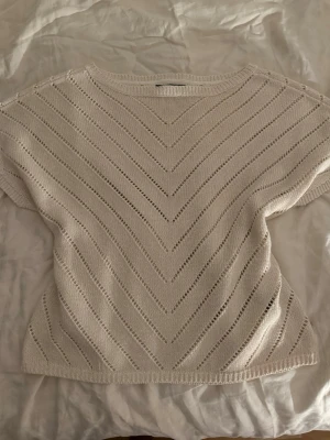 Vit stickad topp med hålmönster - Snygg vit stickad topp med korta ärmar och V-formade hålmönster över hela plagget. Rund halsringning och ribbade kanter. Perfekt att styla med jeans eller kjol och är även lite glittrig 