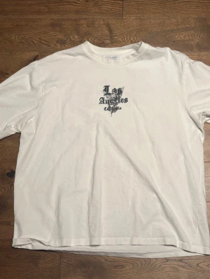Vit t-shirt med Los Angeles-tryck - Säljer en vit t-shirt med svart tryck på bröstet och stort motiv på ryggen med texten 'Los Angeles California' och en ängel. T-shirten har rund hals och korta ärmar. Perfekt för dig som gillar streetwear vibes.