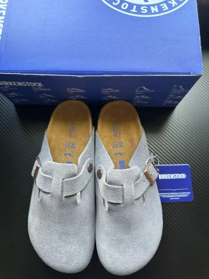 Grå mockatofflor från Birkenstock - Snygga grå tofflor från Birkenstock i mjuk mocka med klassisk slip-in design. De har justerbart spänne i metall och präglad logga på sidan. Fotbädden är i kork och innersulan i mocka för extra komfort. Perfekta för chill dagar hemma eller ute.