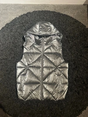 Silvrig dunväst med huva - Snygg silvrig dunväst med dragkedja framtill och stor, vadderad huva. Västen har ett glansigt material och coola diagonala sömmar som ger en modern look. Perfekt för lager på lager och när du vill sticka ut lite extra.