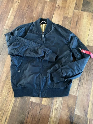 Svart bomberjacka Alpha Industries - Klassisk svart bomberjacka från Alpha Industries med orange innerfoder och ribbade muddar. Jackan har dragkedja framtill, fickor på sidorna och en ikonisk röd tag på ärmen. Tillverkad i slitstarkt nylonmaterial och har en cool militärinspirerad look. Storleken står 2x-large men är mer som L