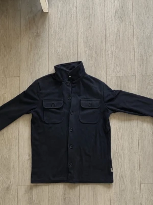 Mörk blå overshirt med fickor - Stilren svart overshirt med knäppning framtill och två bröstfickor med lock. Rak passform och lång ärm, perfekt att bära över en hoodie eller t-shirt. Materialet ser ut att vara mjukt och bekvämt, passar till många olika outfits. Passar en kille runt 175/ 185 cm lång 