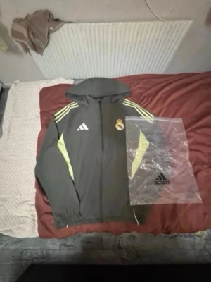 Real Madrid adidas träningsjacka grå - Snygg grå träningsjacka från adidas med Real Madrid-logga på bröstet. Jackan har huva, dragkedja framtill och gula detaljer på ärmarna samt vita adidas-ränder på axlarna. Perfekt för träning eller sportig stil.