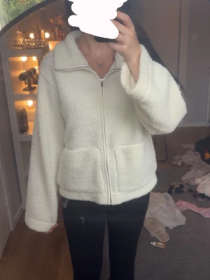 Vit teddyjacka med dragkedja - Mysig vit teddyjacka med dragkedja framtill och två stora fickor. Jackan har en hög krage och är gjord i fluffigt, mjukt material som håller dig varm. Perfekt för kyliga dagar och enkel att matcha med olika outfits.