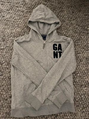Grå zip hoodie från GANT - Snygg grå hoodie från GANT med dragkedja och stor huva. Mjuk bomullsblandning och ribbade muddar vid ärmslut och nederkant. Svart GANT-logga i frotté på bröstet ger en sportig vibe. Perfekt till jeans eller joggers.