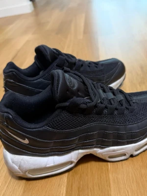 Nike Air Max 95 svarta sneakers - Nike Air Max 95 sneakers i svart med vit sula och synliga Air-enheter. Ovandelen är i mesh och syntet med klassiska vågade paneler och små vita Swoosh-loggor på sidan och hälen. Skorna har snörning och en chunky siluett som ger en sportig vibe.
