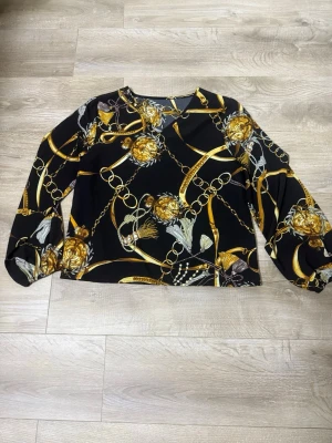 Svart blus med kedjemönster - Trendig svart blus med långa ärmar och ett iögonfallande mönster av guldfärgade kedjor och detaljer. Blusen har en rak passform och rund halsringning. Perfekt för dig som vill sticka ut med en cool och elegant look.