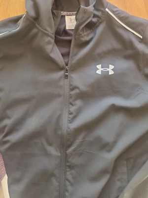 Svart vindjacka från Under Armour - Svart vindjacka från Under Armour med huva och dragkedja framtill. Jackan har vita logotyper på bröstet och ryggen samt diskreta reflexdetaljer vid axlarna. Tillverkad i lätt och vattenavvisande material, perfekt för blåsiga dagar.