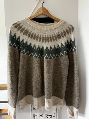 Beige och brun stickad tröja Vero Moda - Stickad tröja från Vero Moda i beige och bruna toner med grönt och vitt mönster över bröstet och axlarna. Tröjan har rund halsringning, långa ärmar och ribbade muddar vid ärmslut och nederkant. Perfekt för kalla dagar och riktigt mysig.