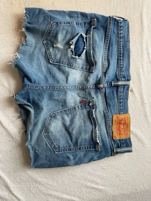 Levi's blå jeansshorts med rå kant - Säljer ett par klassiska blå jeansshorts från Levi's med råa, fransiga kanter och slitningar på bakfickan. Shortsen har fem fickor, knappgylf och den ikoniska Levi's-lappen bak i midjan. Perfekta för sommaren och ger en avslappnad vibe.