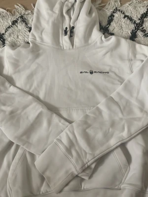 Vit hoodie från Sail Racing - Snygg vit hoodie från Sail Racing med svart logga på bröstet och en unik detalj på huvan. Hoodien har dragsko i huvan och en stor magficka. Tillverkad i mjukt material som känns skönt mot huden. Perfekt för en avslappnad och sportig stil.