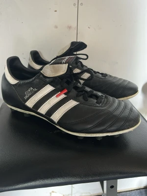 Adidas Copa Mundial fotbollsskor svart/vit - Klassiska Adidas Copa Mundial fotbollsskor i svart läder med vita detaljer och de ikoniska tre ränderna på sidan. Skorna har snörning, låg profil och dobbar för spel på gräs. Perfekta för dig som vill ha en tidlös och bekväm sko på planen.