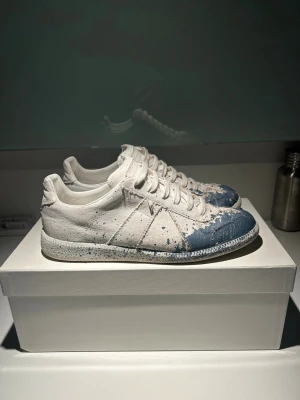 Vita med blå paint splatter Maison Margiela - Unika skor i vitt tyg med blå färgdetaljer och coola färgstänk över hela skon. Lågt skaft och klassisk snörning. Snygg kontrast mellan det vita och blåa, perfekt för dig som vill sticka ut med din stil. Äkta Storlek 40 MEN 41 om man tar av sulan
