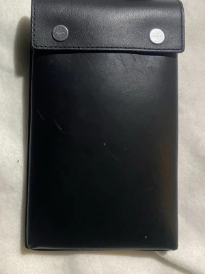 Svart axelväska i läder med spänne - Stilren svart axelväska i slätt läder med lock och två silverfärgade knappar. Justerbar axelrem i nylon med spänne och metallring. Perfekt för att bära det viktigaste och passar till många outfits.