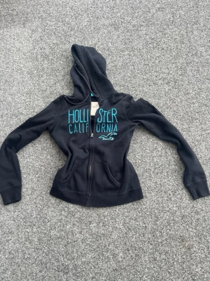 Svart Hollister hoodie med turkos text - Snygg svart hoodie från Hollister med dragkedja och turkos broderad text 'Hollister California' på bröstet. Klassisk huva och två fickor framtill. Tillverkad i mjukt bomullsmaterial, perfekt för chill dagar eller när du vill ha en avslappnad look.